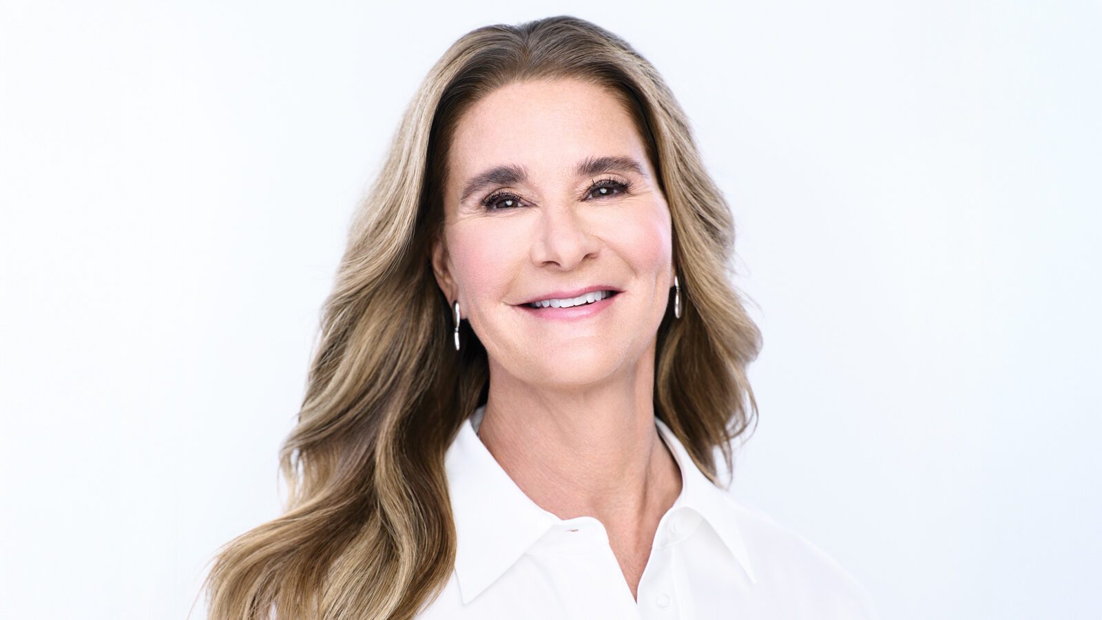 A woman with long, wavy brown hair and light skin smiles at the camera. She is wearing a white blouse and small drop earrings, embodying the grace often associated with Melinda French Gates of the Gates Foundation, against a plain white background.