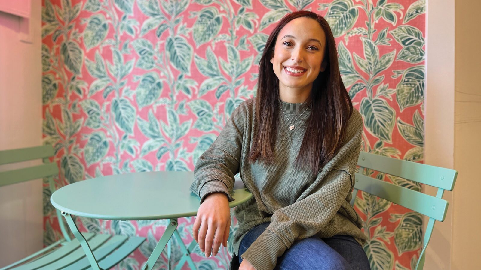 Rachel Barnecut sits at a mint green table and chair set in front of pink and green leaf-patterned wallpaper, smiling at the camera—a fitting portrait among trailblazing women.