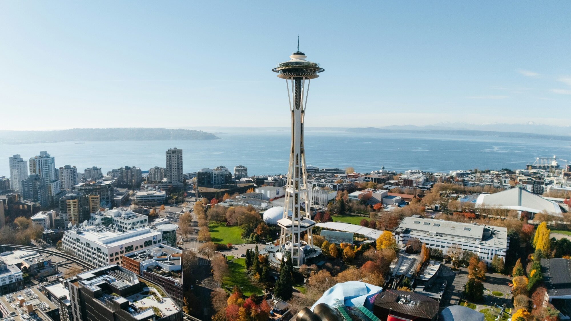 Aerial view of Seattle, Washington, featuring the Space Needle in the center, surrounded by buildings, trees, and water in the background under a clear sky.