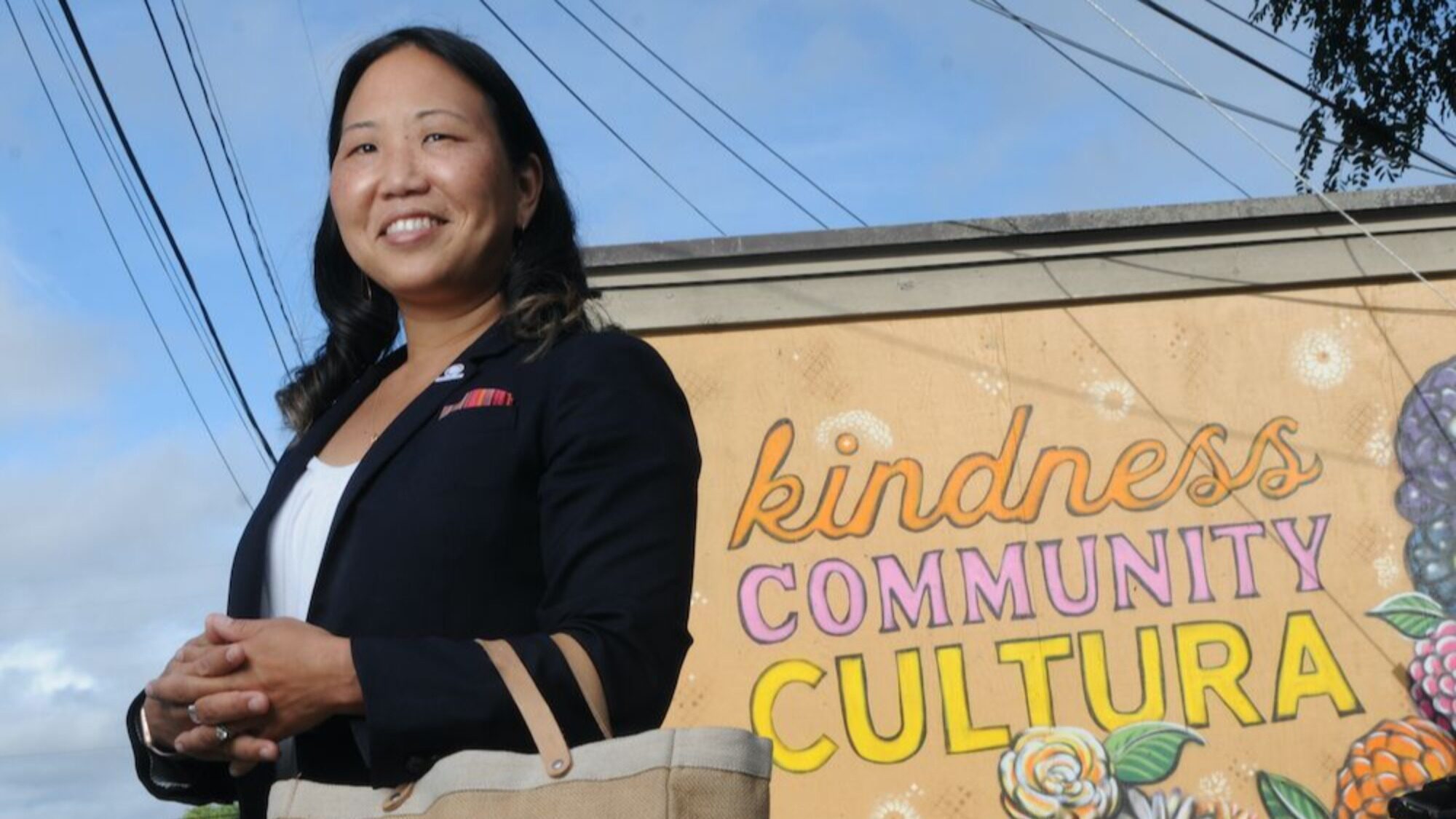 A person stands in front of a mural with the words "kindness COMMUNITY CULTURA" and floral designs, holding a beige bag and smiling.