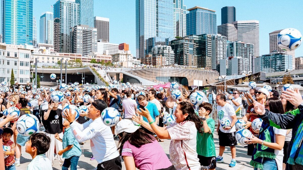 A large crowd outdoors in a city catches or tosses blue and white soccer balls, with tall buildings and clear skies in the background.