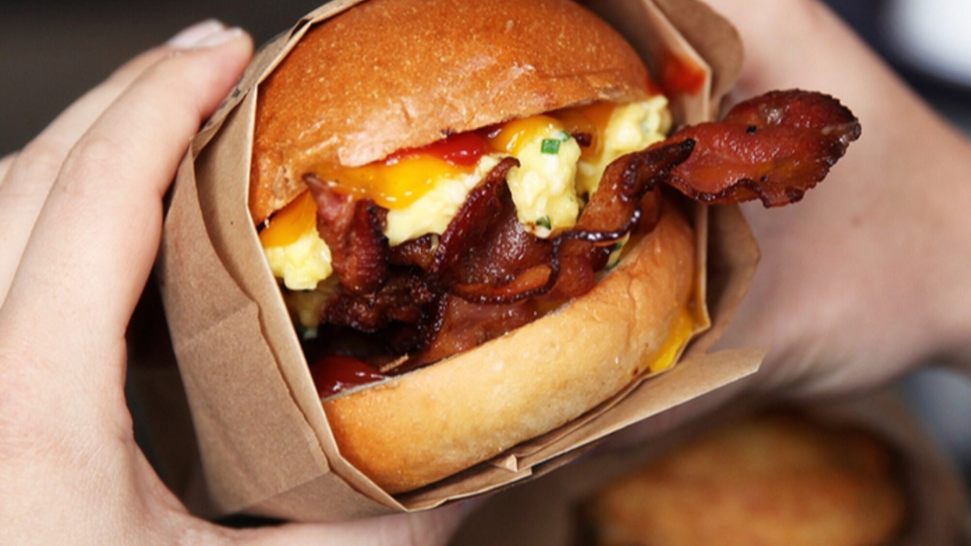 A person holds a sandwich with crispy bacon, scrambled eggs, melted cheese, and sauce in a brown paper wrapper.