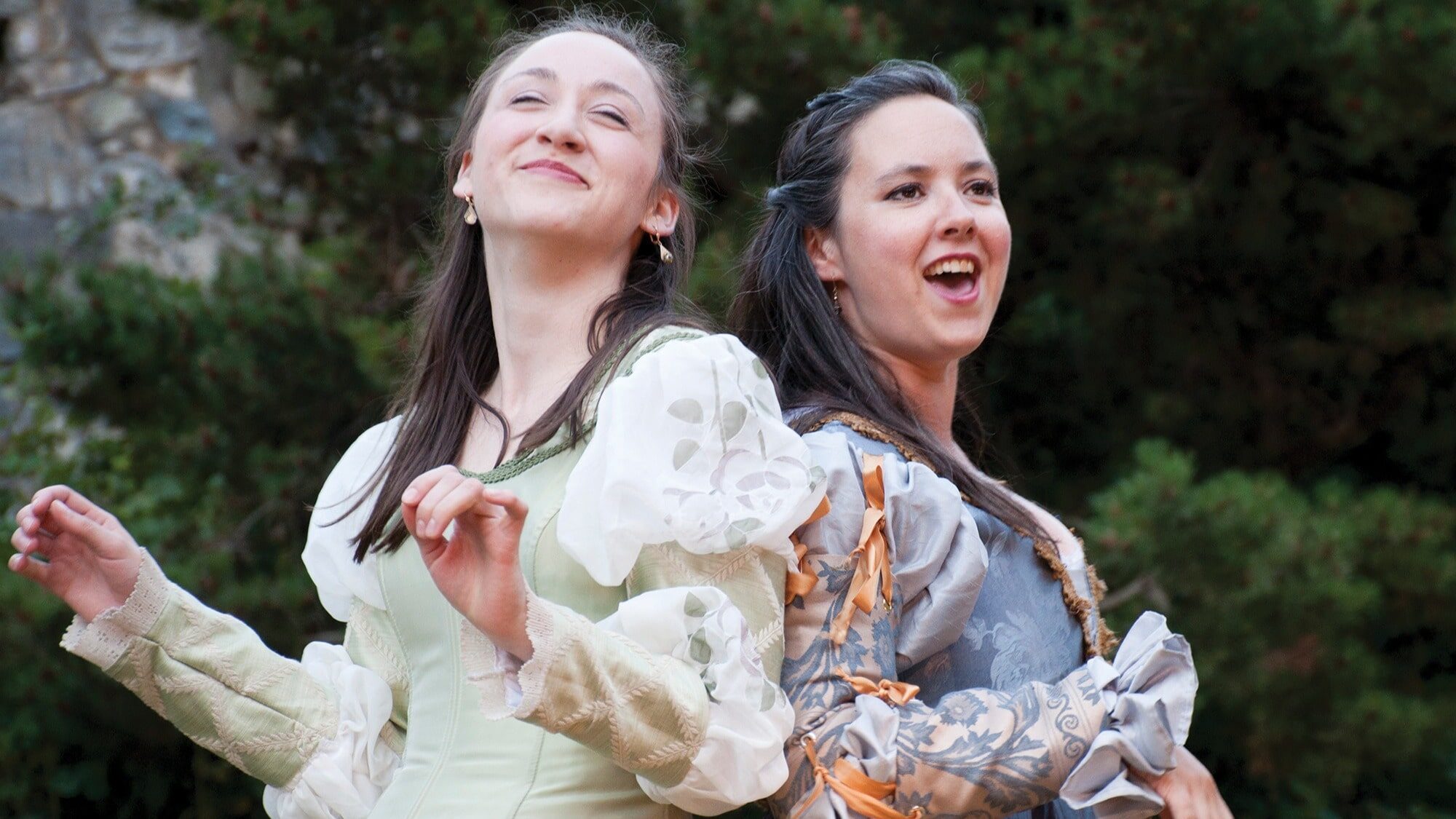 Two women in historical costumes stand back to back outdoors, smiling and gesturing with their hands at a lively food festival, surrounded by greenery in the background.