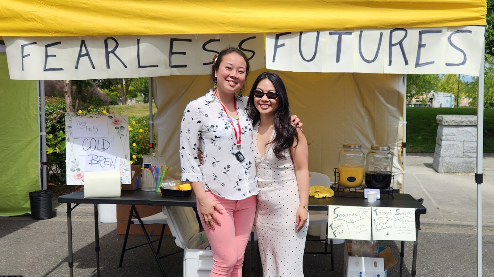 Two women stand smiling in front of a coffee stand with a "Fearless Futures" banner, cold drinks, and a handwritten menu at an outdoor event, welcoming everyone.