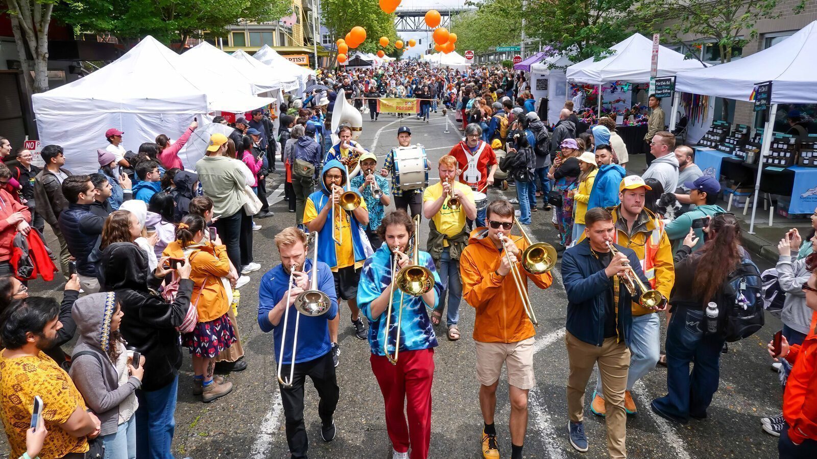 A brass band marches through a crowded outdoor street festival lined with vendor tents, as people watch and take photos—one of the many fun things to do in Seattle near MOPOP and SeattleRep.