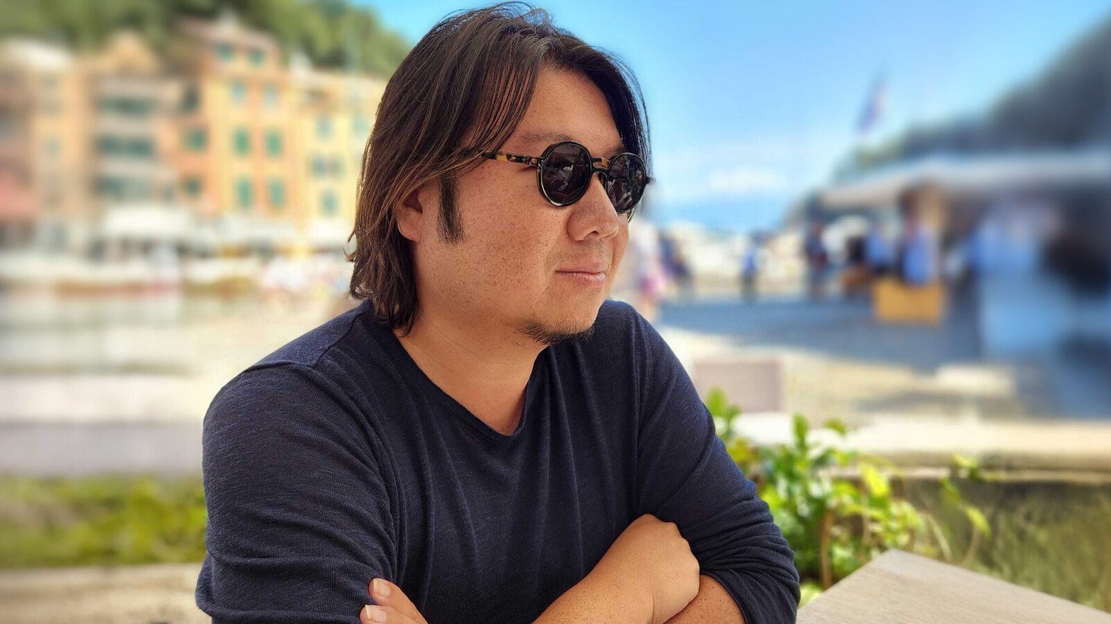 A man with medium-length hair and sunglasses sits outdoors at a table with his arms crossed, evoking the stylish flair of Crazy Rich Asians, with a blurred background of buildings and people.