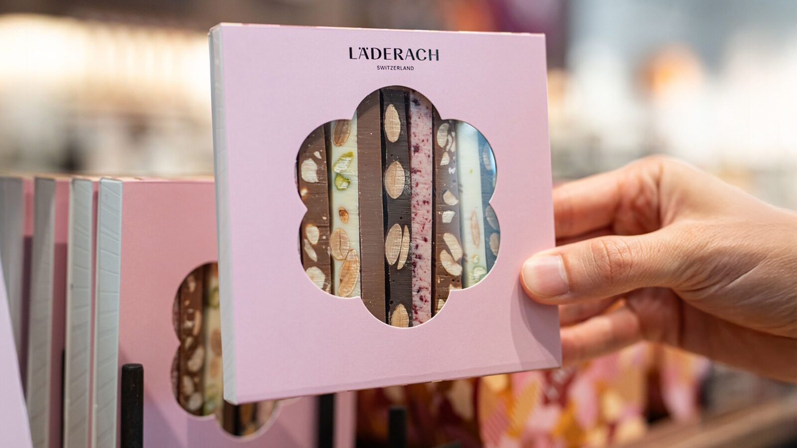 A hand holds a pink box of Läderach Swiss chocolate bars, fresh from Switzerland, featuring a flower-shaped window that reveals assorted flavors with nuts.
