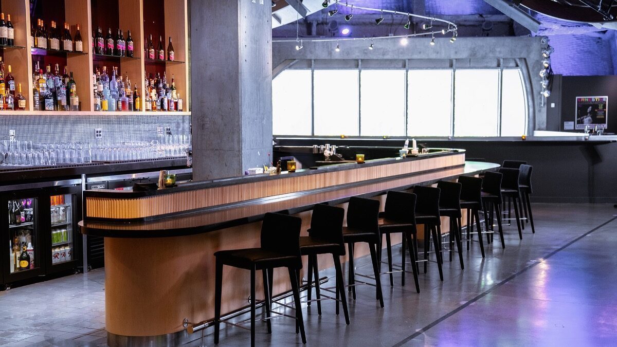 Modern bar interior with a long wooden counter, black bar stools, shelves stocked with bottles, and large windows in the background.