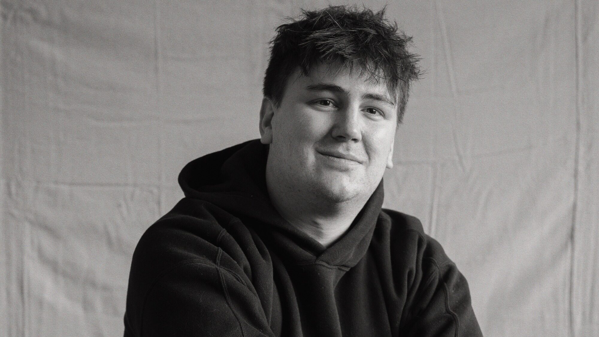 A young man with short, tousled hair wearing a dark hoodie sits in front of a plain cloth backdrop, looking at the camera and smiling slightly.