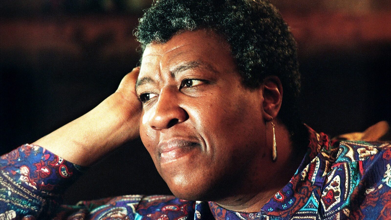 A person with short curly hair, wearing a patterned shirt and earring, rests their head on their hand and looks thoughtfully into the distance, as if ready to celebrate Octavia E. Butler Day and her enduring legacy.