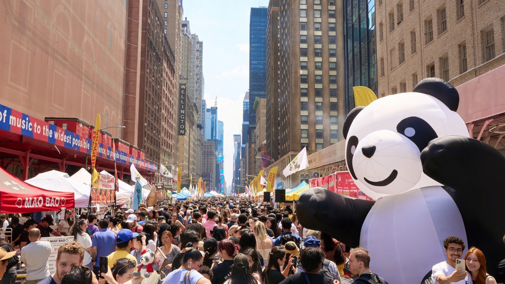 A large crowd fills a city street lined with vendor booths, with a giant inflatable panda and tall buildings visible in the background.
