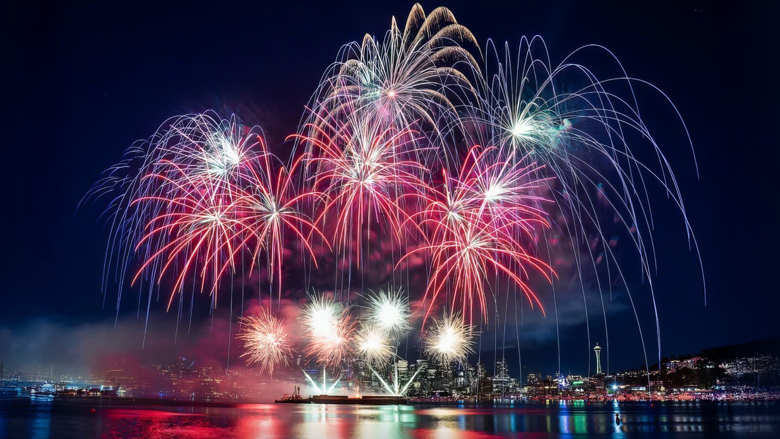 Fireworks in red, white, and blue burst over a city skyline at night, reflecting on the water below—a scene worthy of Lit Up: Five Fireworks Shows That Dazzle the Planet.
