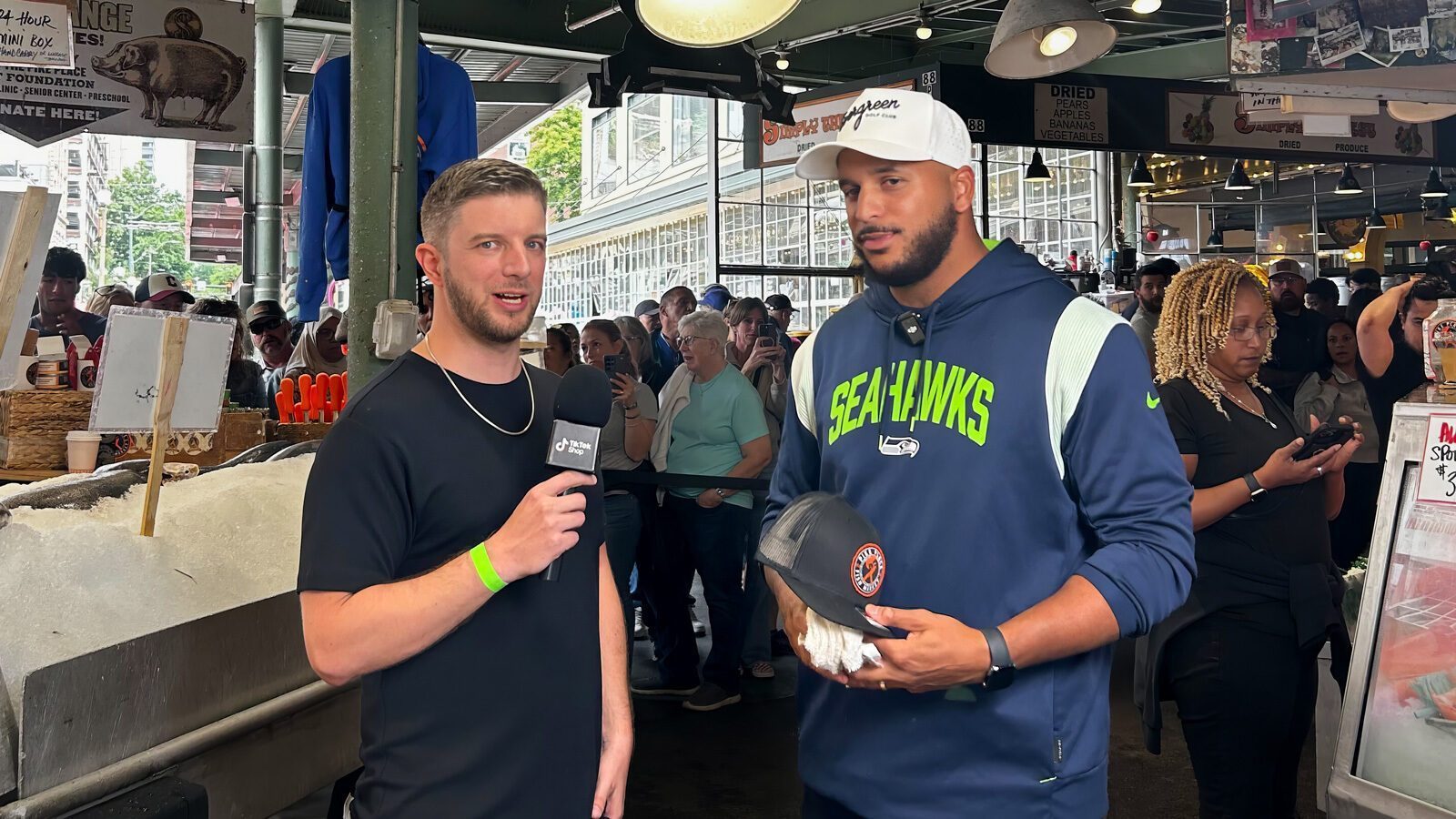 Two men stand in a busy market; one holds a microphone and speaks, while the other wears a Seahawks hoodie and holds a wrapped item. People and market stalls are visible in the background.