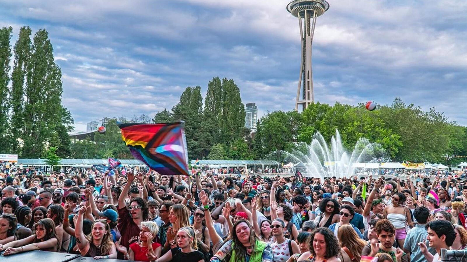 A large crowd gathers at an outdoor event near a fountain, with a Progress Pride flag visible and the Space Needle in the background on a partly cloudy day as vibrant celebrations honor Seattle’s rich LGBTQIA+ history.