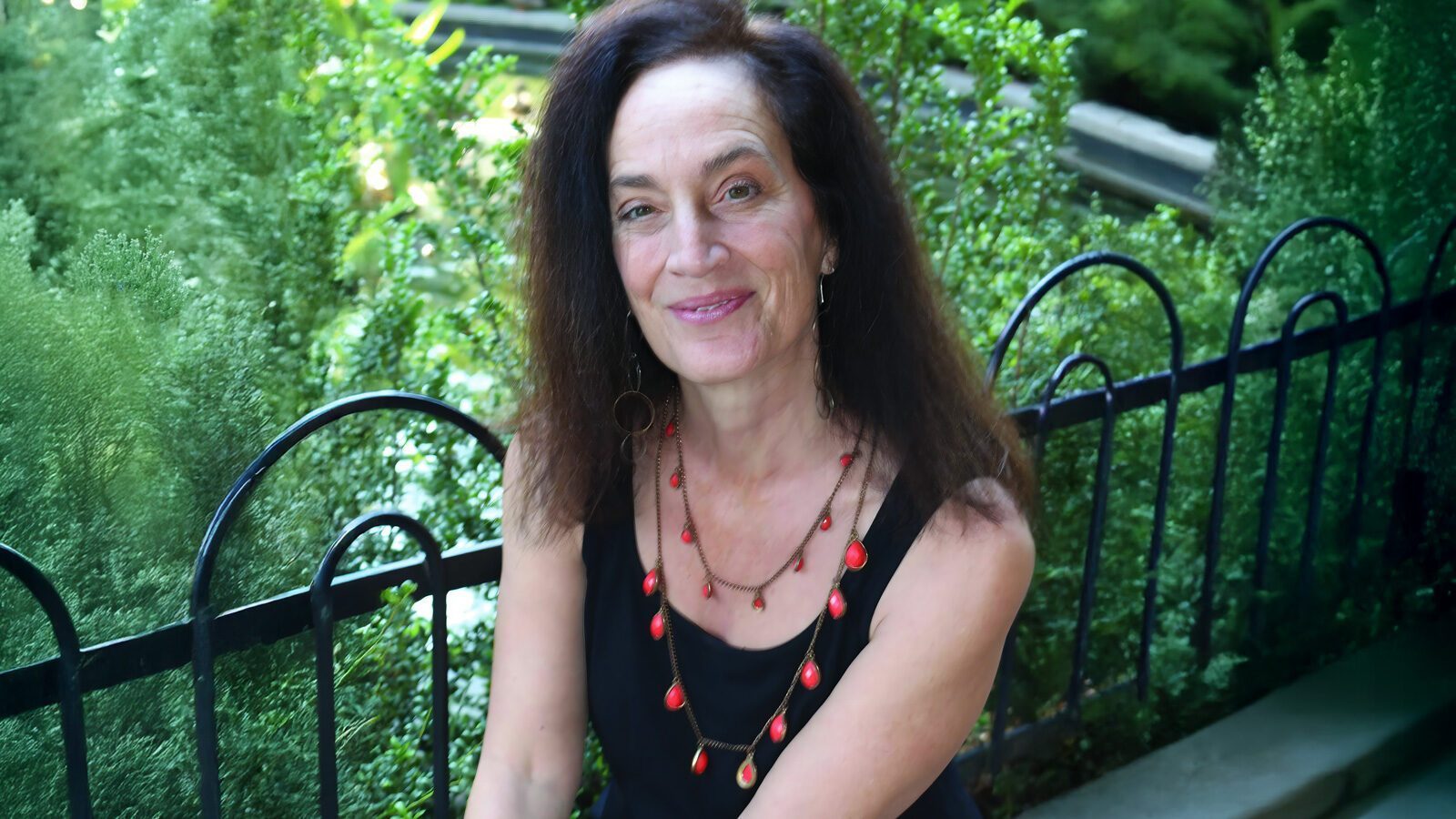 Ann Muno sits in front of green bushes and a black metal fence, her long dark hair flowing over a black sleeveless top and red bead necklace—dedicated to raising compassionate young women through the Justice for Girls Coalition Washington State.