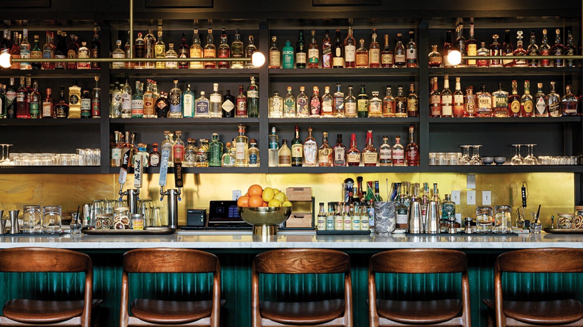 A bar interior at Ramie, one of the standout Capitol Hill restaurants, features liquor bottles on shelves, a countertop with bar tools and fruit, and four wooden bar stools inviting guests to enjoy Trinh Nguyen’s signature creations.