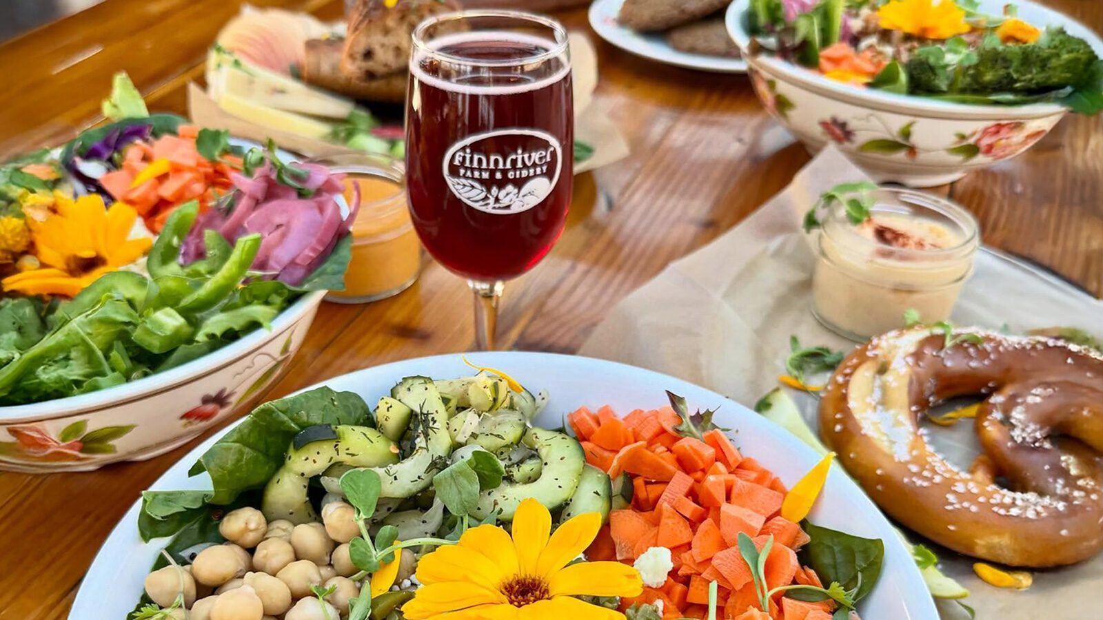 A table set with colorful salads, a pretzel, dips, and a glass of Finnriver Farm & Cidery’s red cider, garnished with edible flowers—a vibrant spread perfect for food delivery or enjoying fresh.