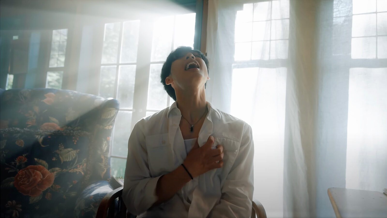 A person sits indoors with sunlight streaming through windows, head tilted back and hand placed on their chest, wearing a white shirt and necklace—capturing a romantic mood reminiscent of a scene from Johnny Huynh’s new music video.