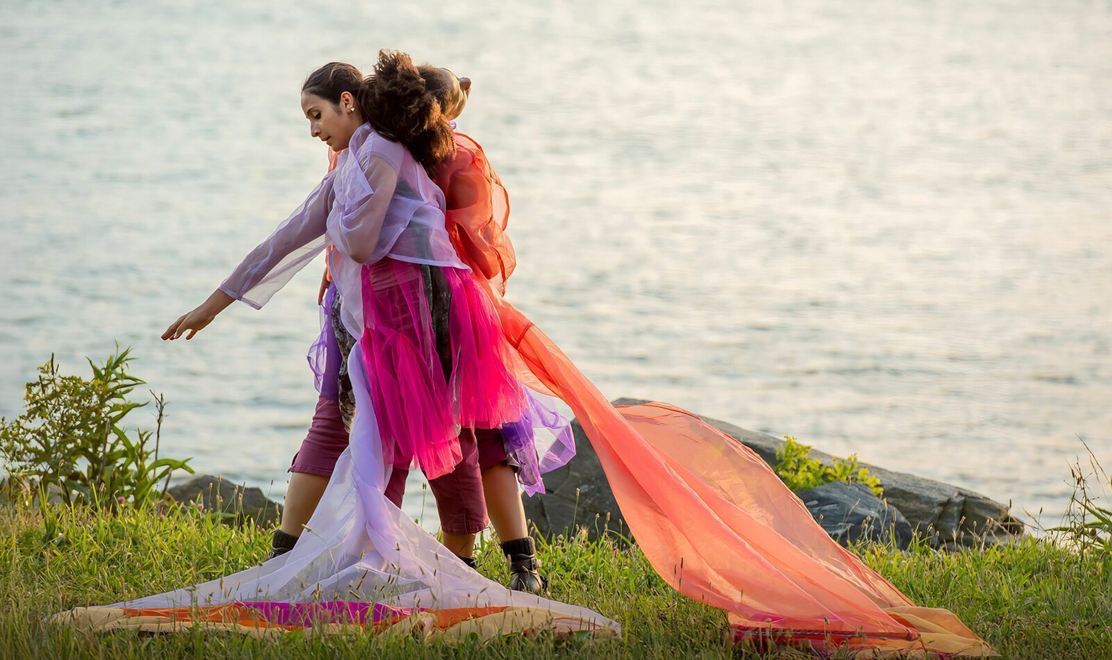 Two people wearing colorful, translucent fabric walk on grass near a body of water, with long pieces of fabric trailing behind them.