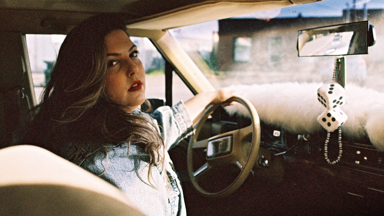A woman with long hair sits in the driver's seat of a car, looking back over her shoulder as if she's finding her way back. Fuzzy dice and beads hang from the rearview mirror.