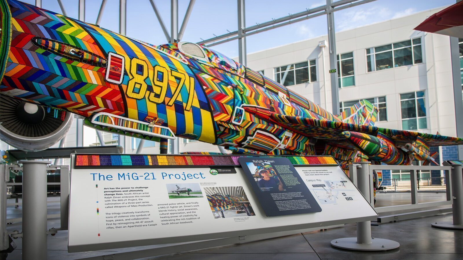 A MiG-21 jet, symbolizing both war and art, is painted in bright, multicolored geometric patterns and displayed in a museum with an informational sign about the MiG-21 Project in front.
