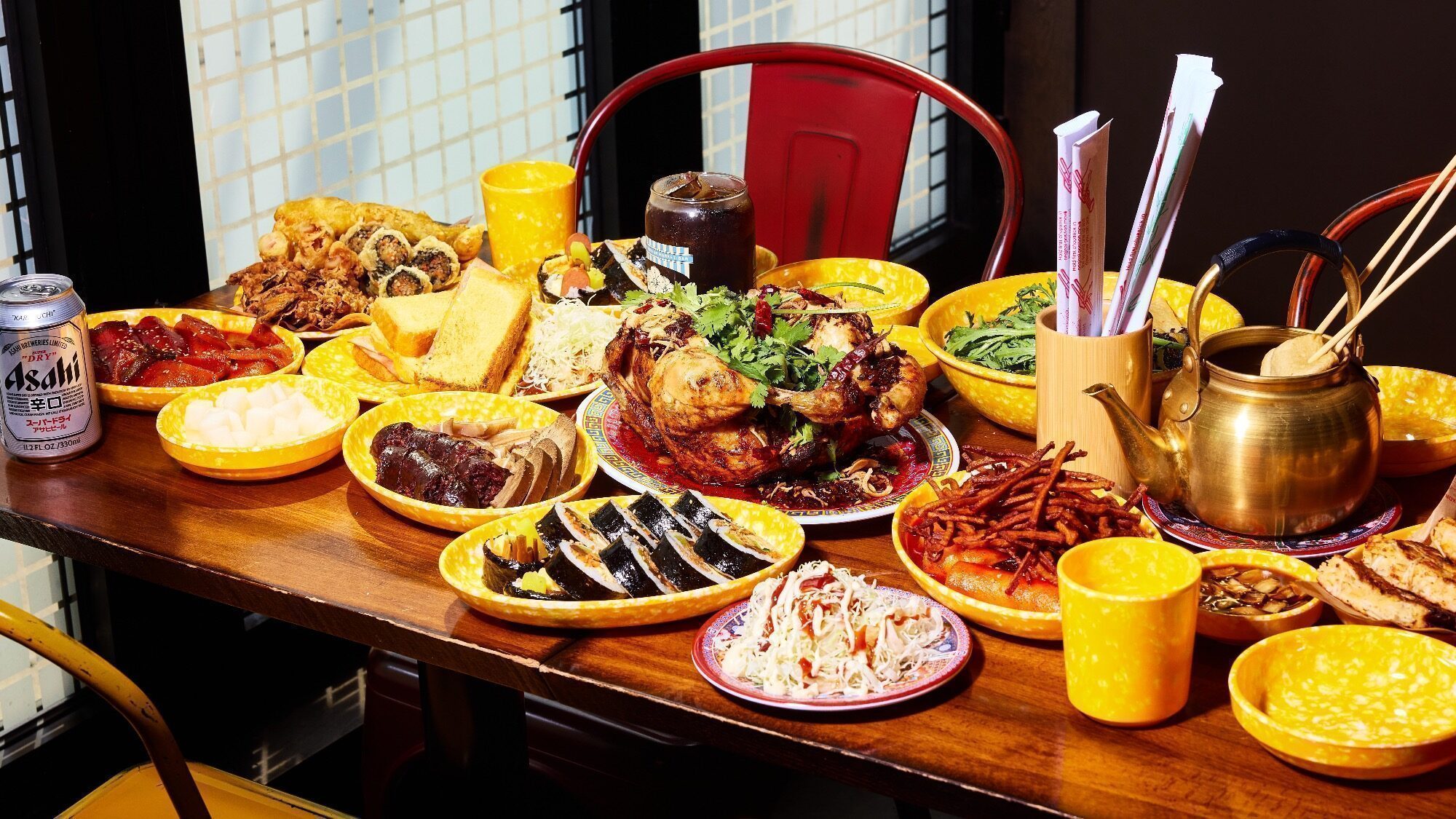 A wooden table set with various Korean dishes, including kimbap, fried chicken, noodles, pickles, side dishes, and drinks, surrounded by yellow plates and utensils.