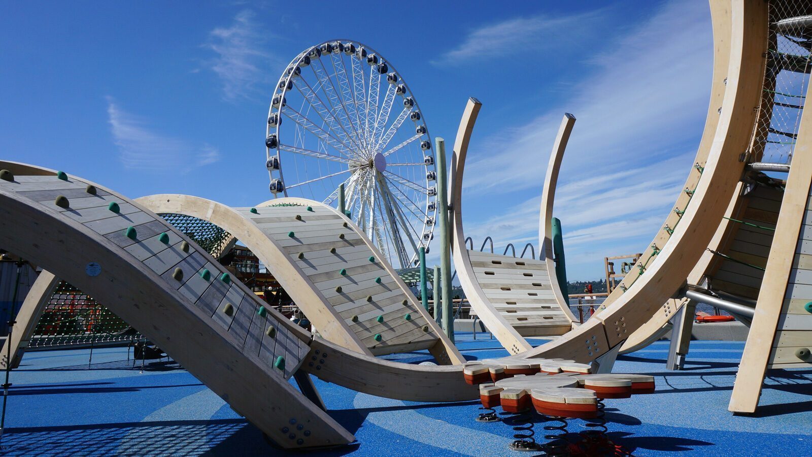 A playground with curved wooden climbing structures and a blue rubber surface invites Playtime on the Pier, with a large Ferris wheel in the background under a clear sky.