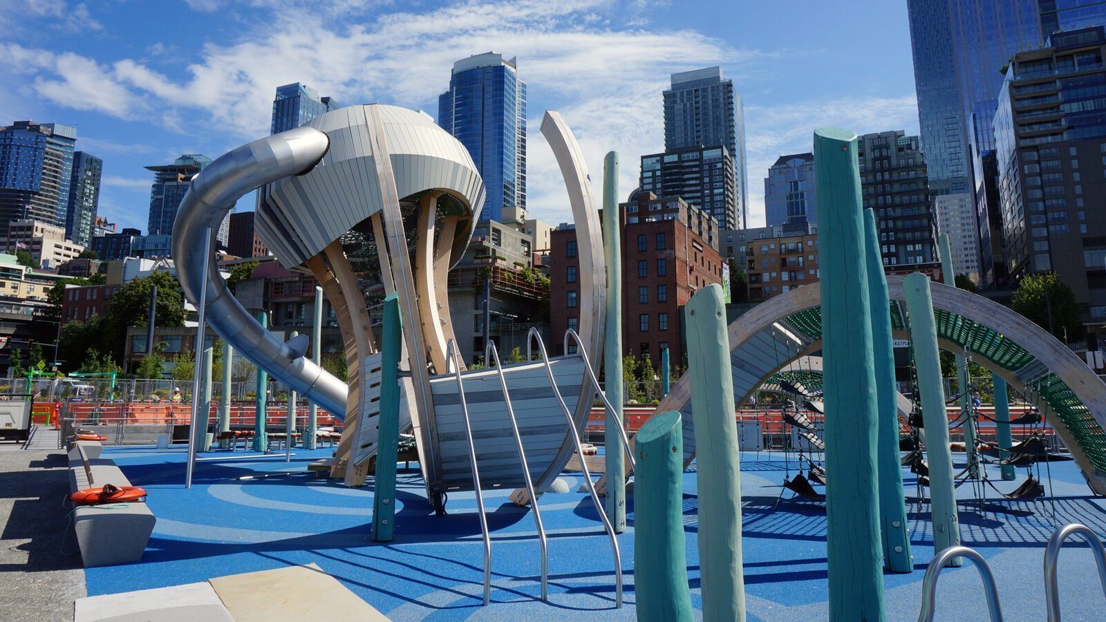 Modern urban playground with climbing structures and slides offers endless playtime fun, set against a backdrop of tall buildings and a blue sky near the lively pier.