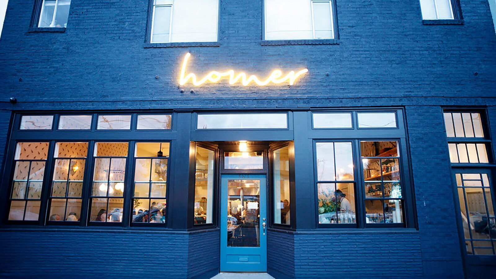 Exterior of a dark blue brick restaurant building with large windows and a lit sign above the door reading “homer”; indoor lighting and diners visible inside, welcoming guests for the Homer Reopening featured in Restaurant Roundup.