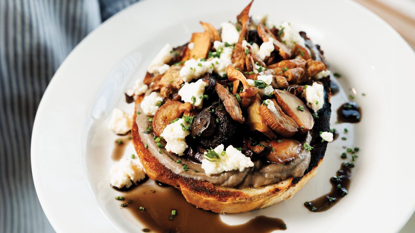 A toasted bread slice topped with sautéed mushrooms, crumbled cheese, and chopped chives, served with a dark sauce on a white plate at Robin's Restaurant and Market—one of the standout new restaurants in Seattle.