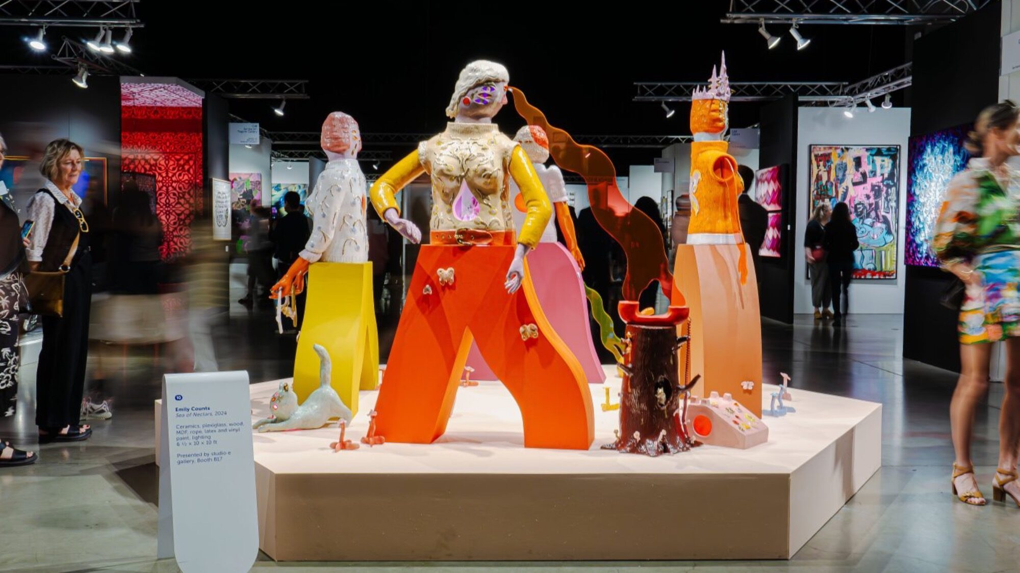 A colorful art installation featuring abstract human-like figures with bright, bold shapes and surreal details is displayed in a gallery with people viewing the exhibit.