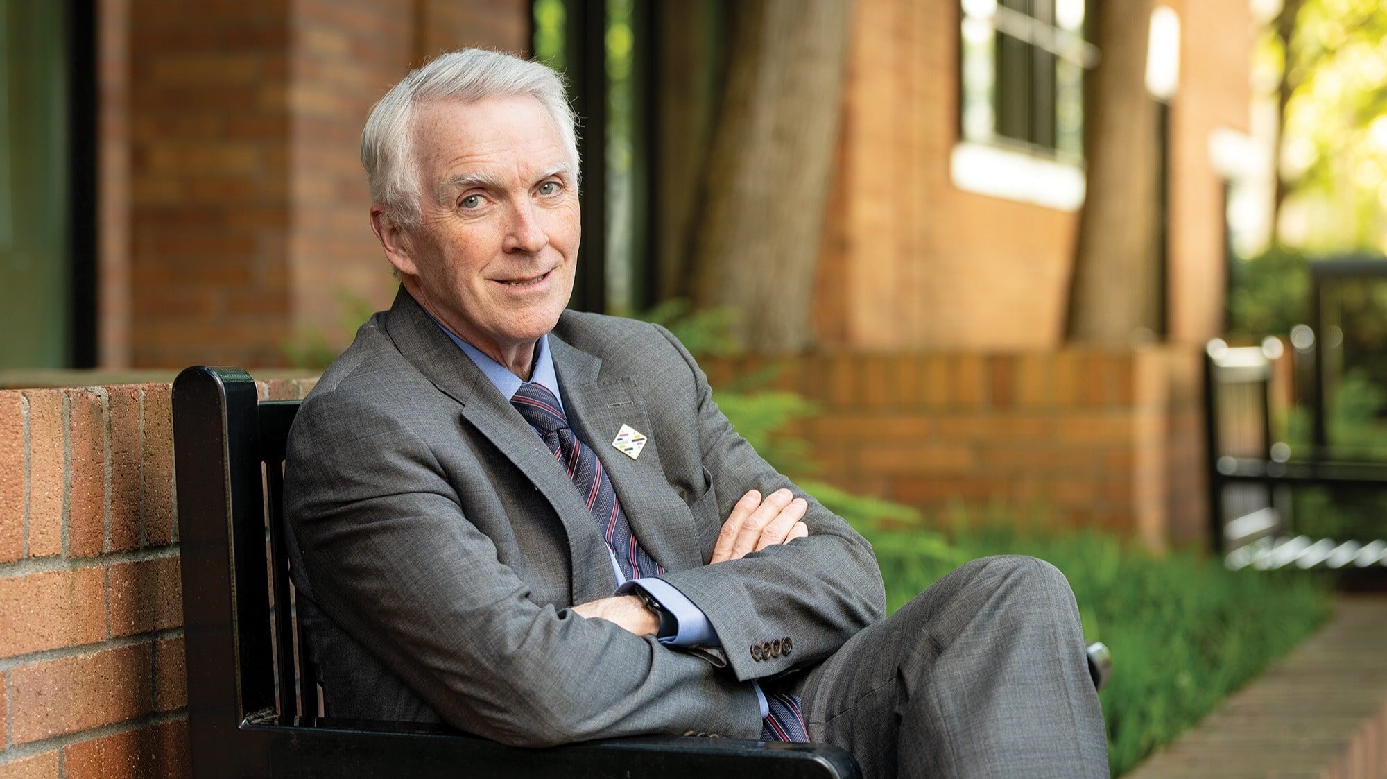 An older man in a gray suit sits on an outdoor bench with his arms crossed, smiling slightly—a look of quiet victory and vision. Brick building and greenery are visible in the background.