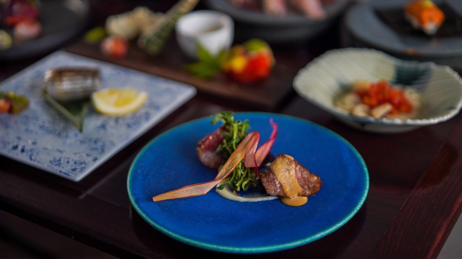 A blue plate with seared meat, sauce, and garnish is in focus; other assorted dishes are blurred in the background on a dark table.