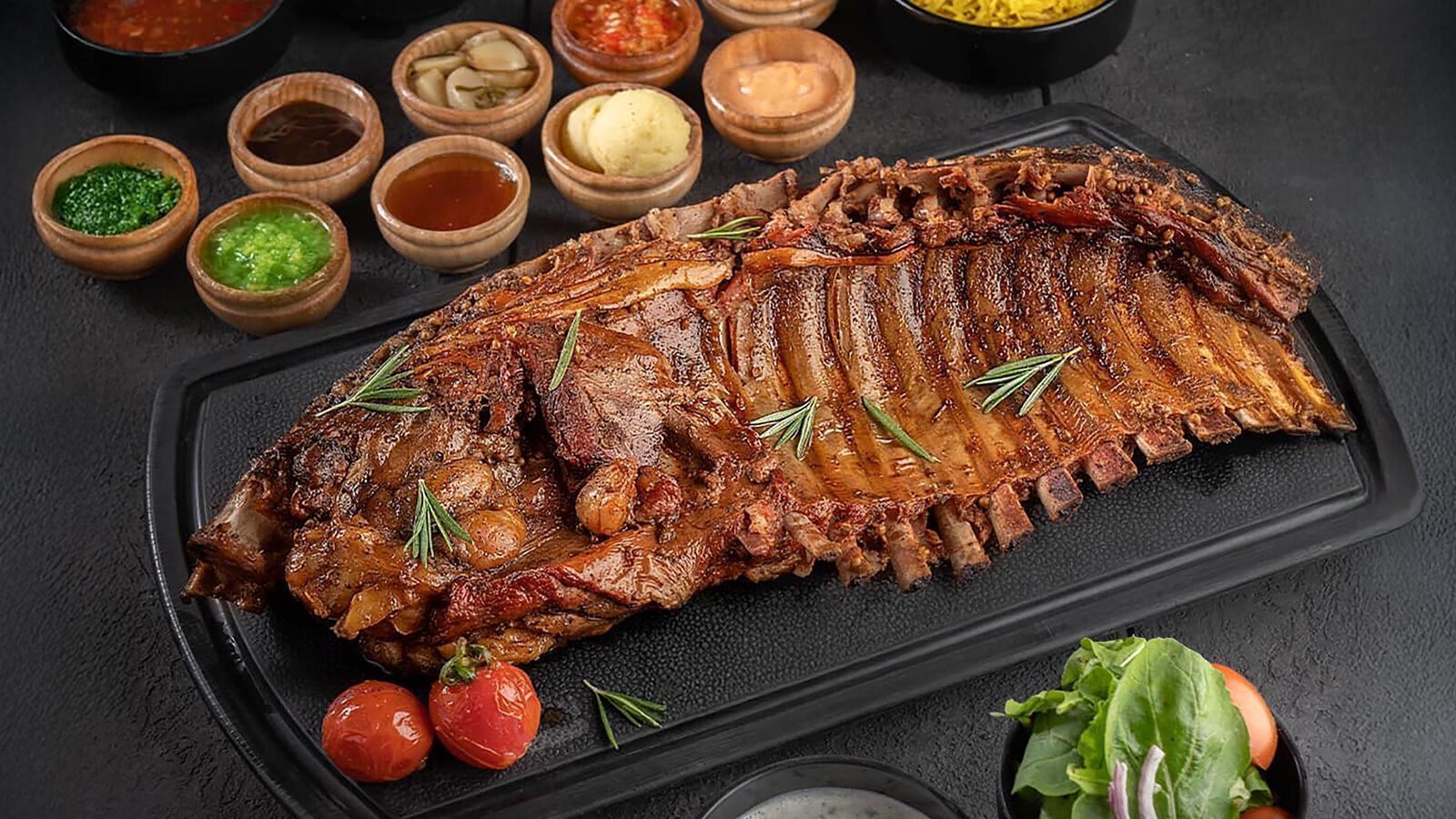 A large cooked rack of ribs from PCC Corner Market is garnished with rosemary and served on a black tray, accompanied by cherry tomatoes, sauces in small bowls, and a side salad—a true high-end halal feast.