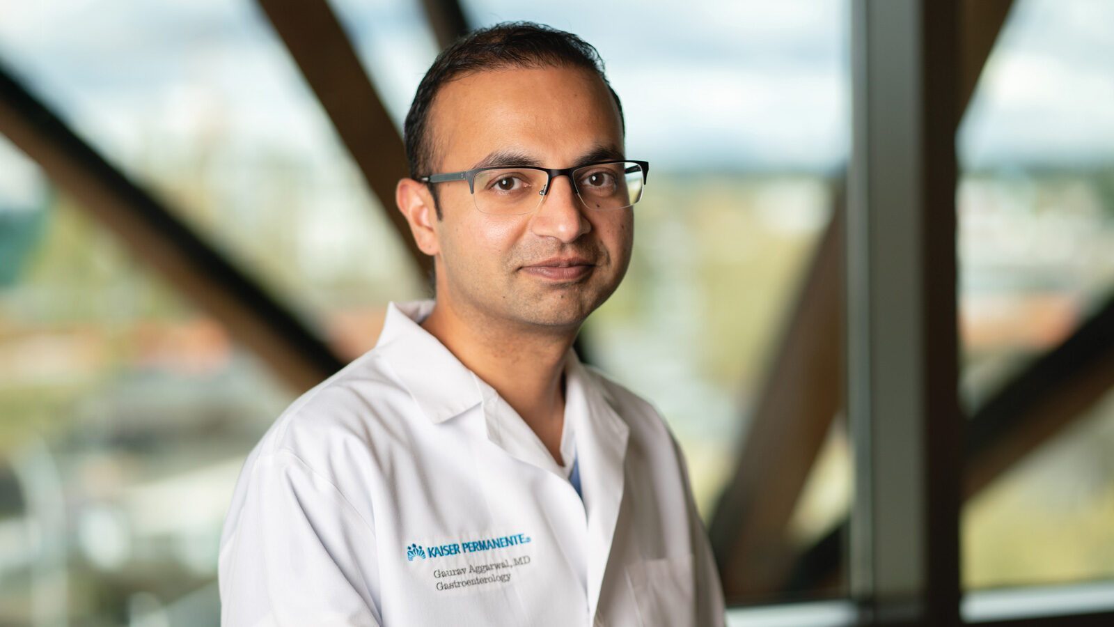 Dr. Gaurav Aggarwal, M.D., in glasses and a white lab coat, stands indoors before large windows with blurred structural beams in the background.