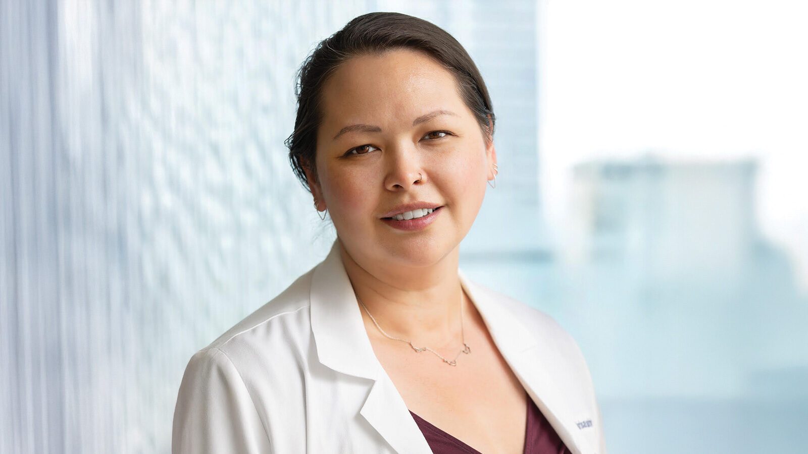 Dr. Marisa Dahlman, M.D., in a white lab coat, stands in front of a window, smiling slightly at the camera with a blurred cityscape in the background.