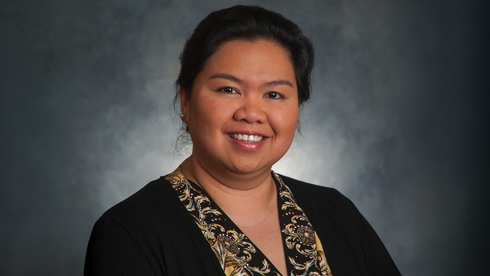 Dr. Mary Ann Pefanco, M.D., with dark hair pulled back and wearing a black top with a patterned collar, smiles at the camera against a dark, blurred background.