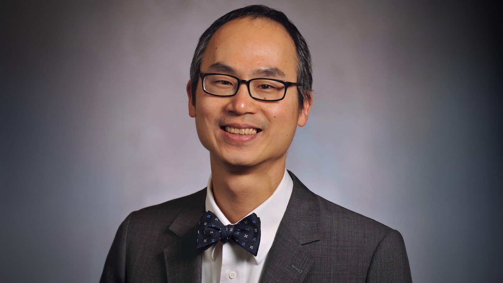Dr. Tobias Lee, M.D., wearing glasses, a dark suit, a white shirt, and a polka dot bow tie, smiles in front of a plain background.