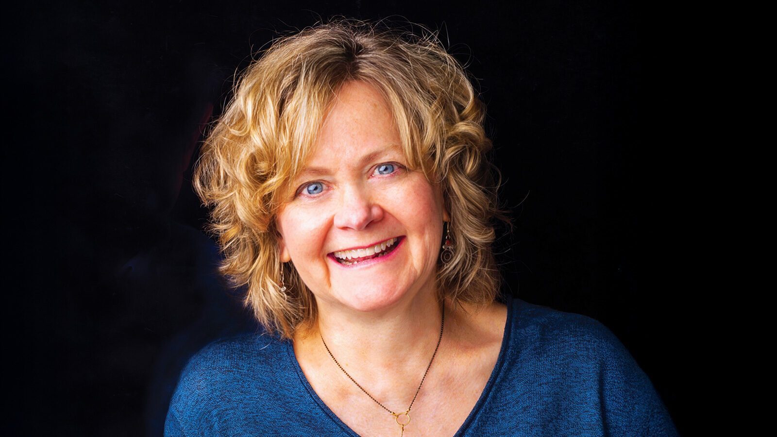 A woman with curly blonde hair and blue eyes smiles at the camera, wearing a blue top and a simple necklace against a black background, embodying the welcoming spirit of Providence Monroe Family Medicine.