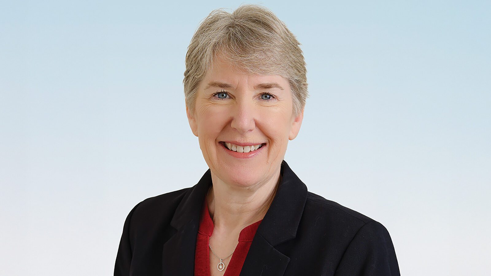 Dr. Kimberly Mebust, with short gray hair, wearing a black blazer over a red top, smiles at the camera against a light blue background.