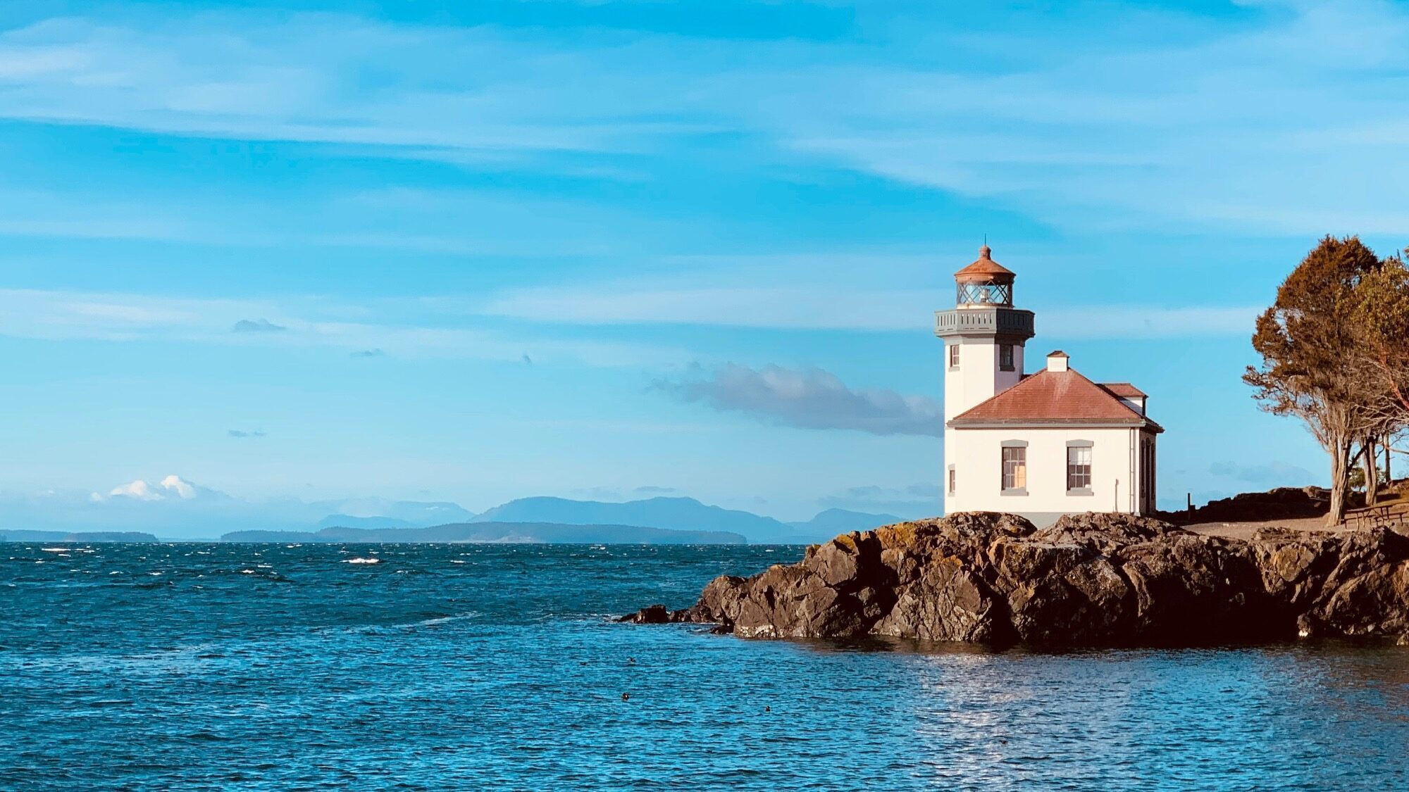 A white lighthouse with a red roof stands on a rocky shoreline, overlooking a blue sea under a clear sky with distant mountains in the background.