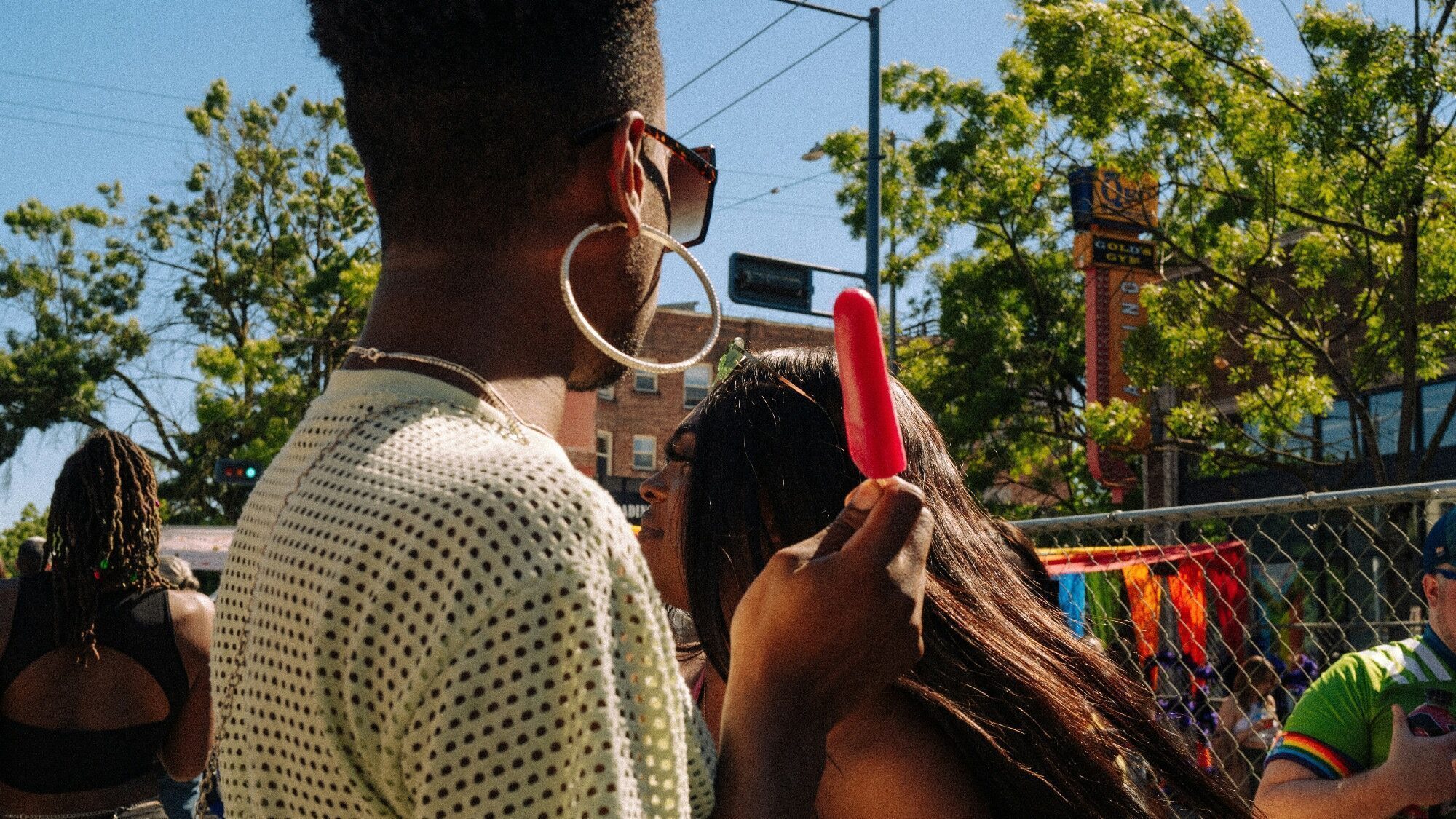 A person wearing sunglasses and large hoop earrings holds a red popsicle at an outdoor event, capturing The Pulse of summer with Bring the Heat vibes; other people and trees are visible in the background.