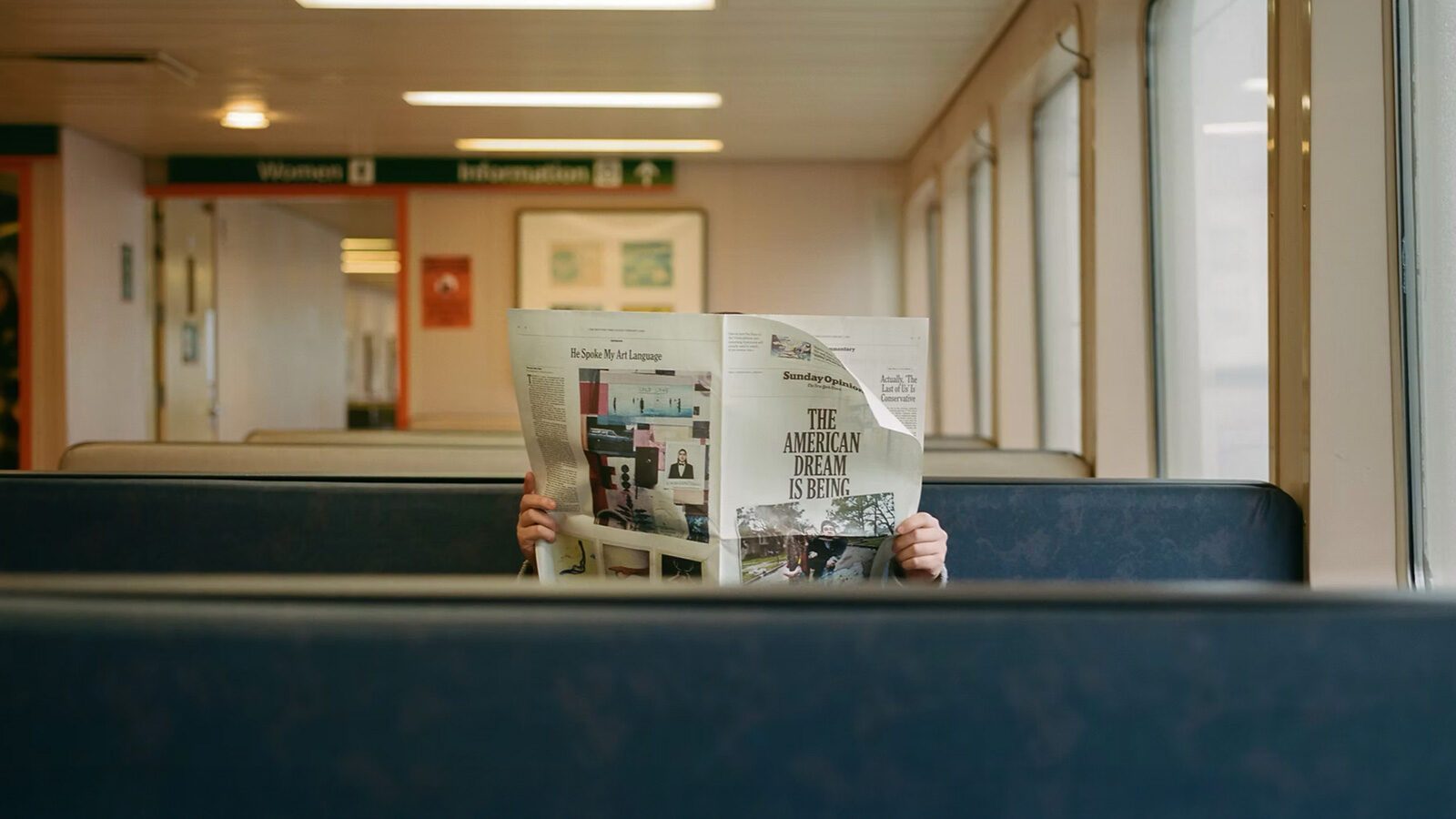 A person sits alone on a Seattle ferries bench, face hidden behind a newspaper titled "The American Dream is Being...," in a candid moment captured by Kristopher Shinn using 35mm photography.