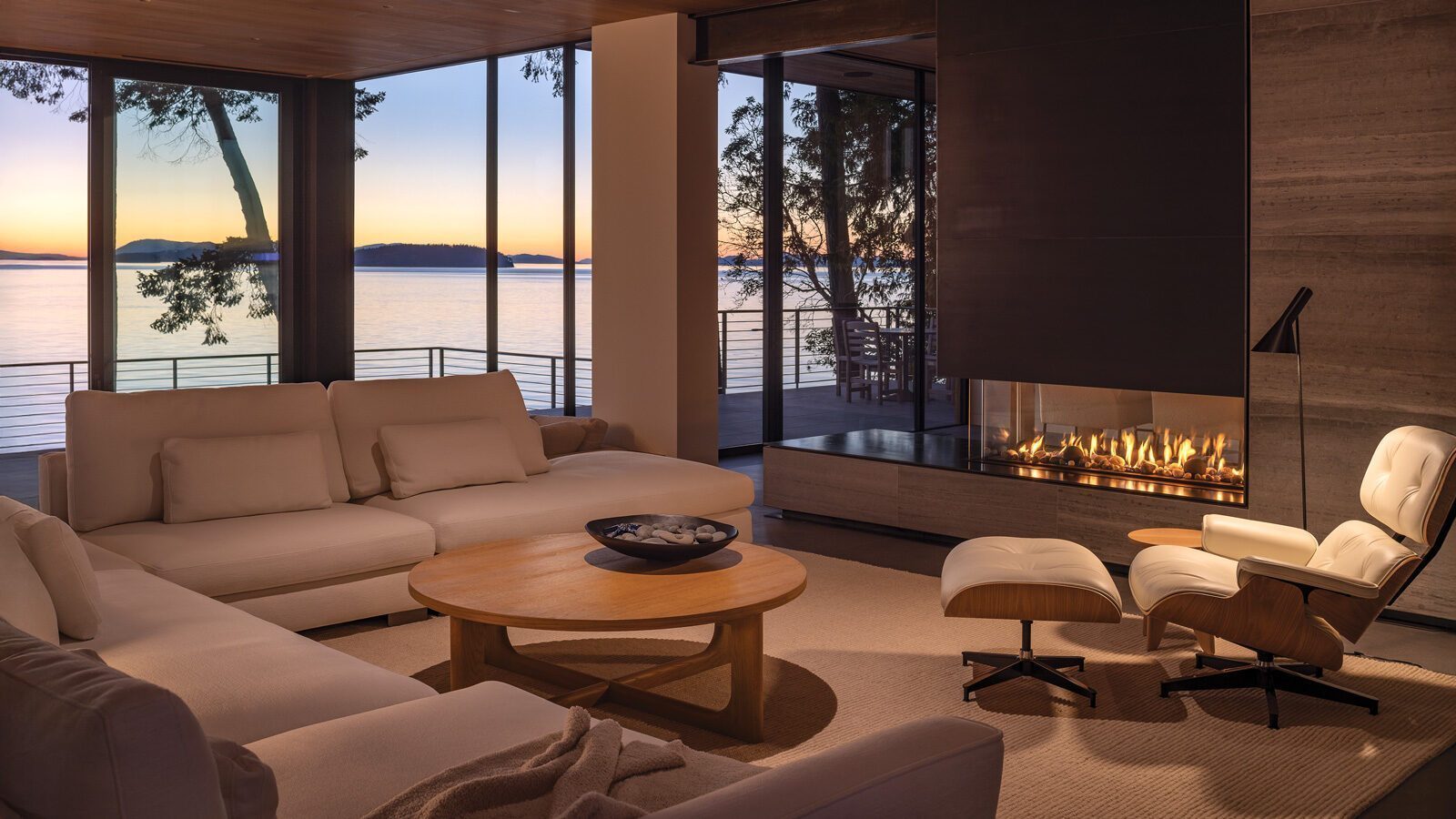 Modern living room with neutral sofas and a lounge chair around a glass fireplace, overlooking the lake at sunset through large windows and sliding doors to the deck—an inviting space inspired by Orcas Island home renovation.