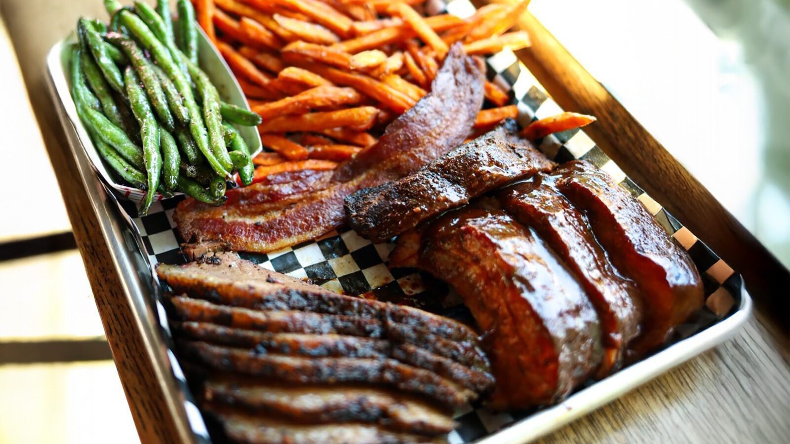 A tray of Brileys BBQ delights featuring sliced brisket, ribs, bacon, sweet potato fries, and green beans served on a checkered paper liner.