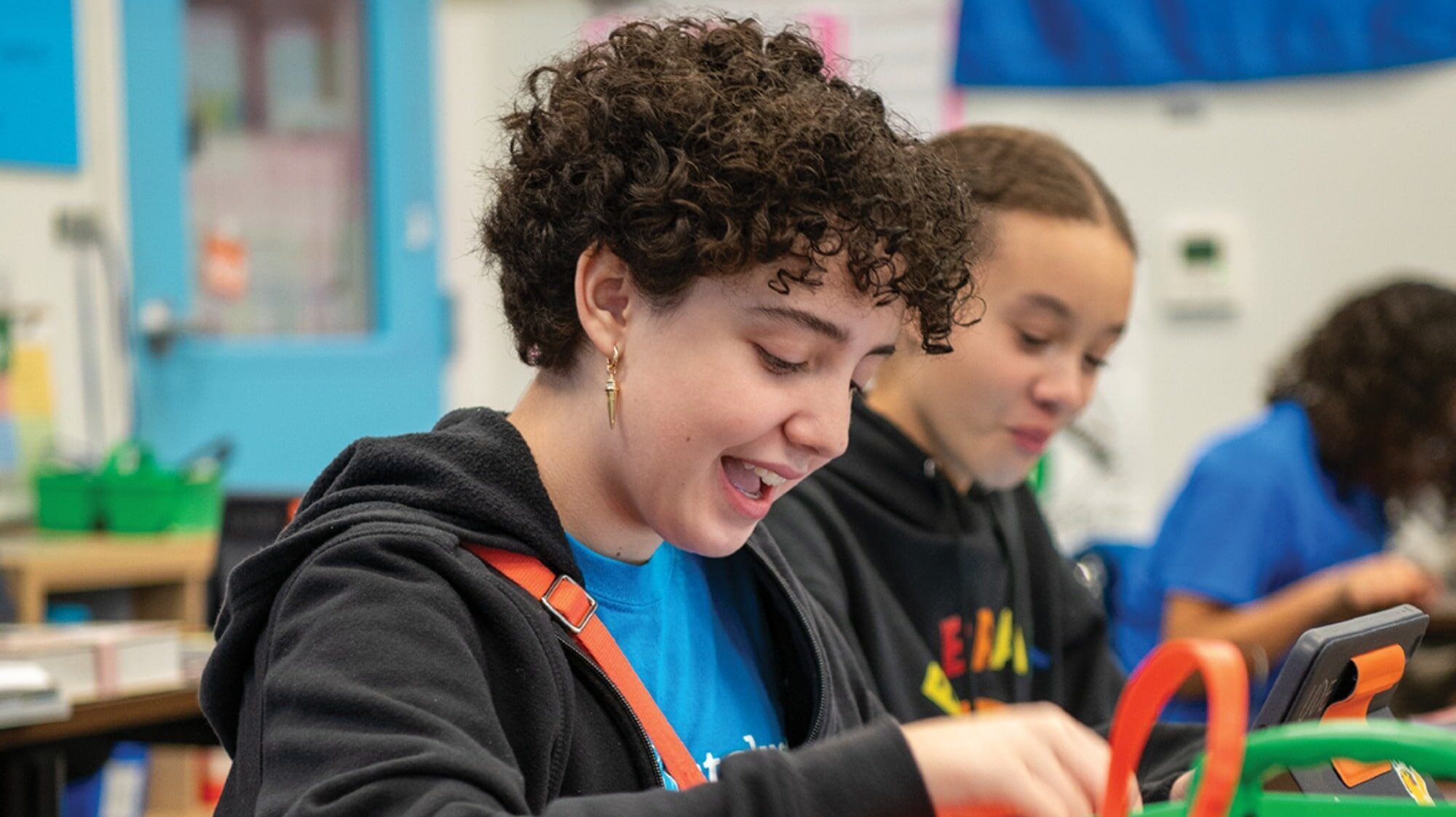 A student with curly hair smiles while working at a desk in a classroom, considering more options, with another student in the background.