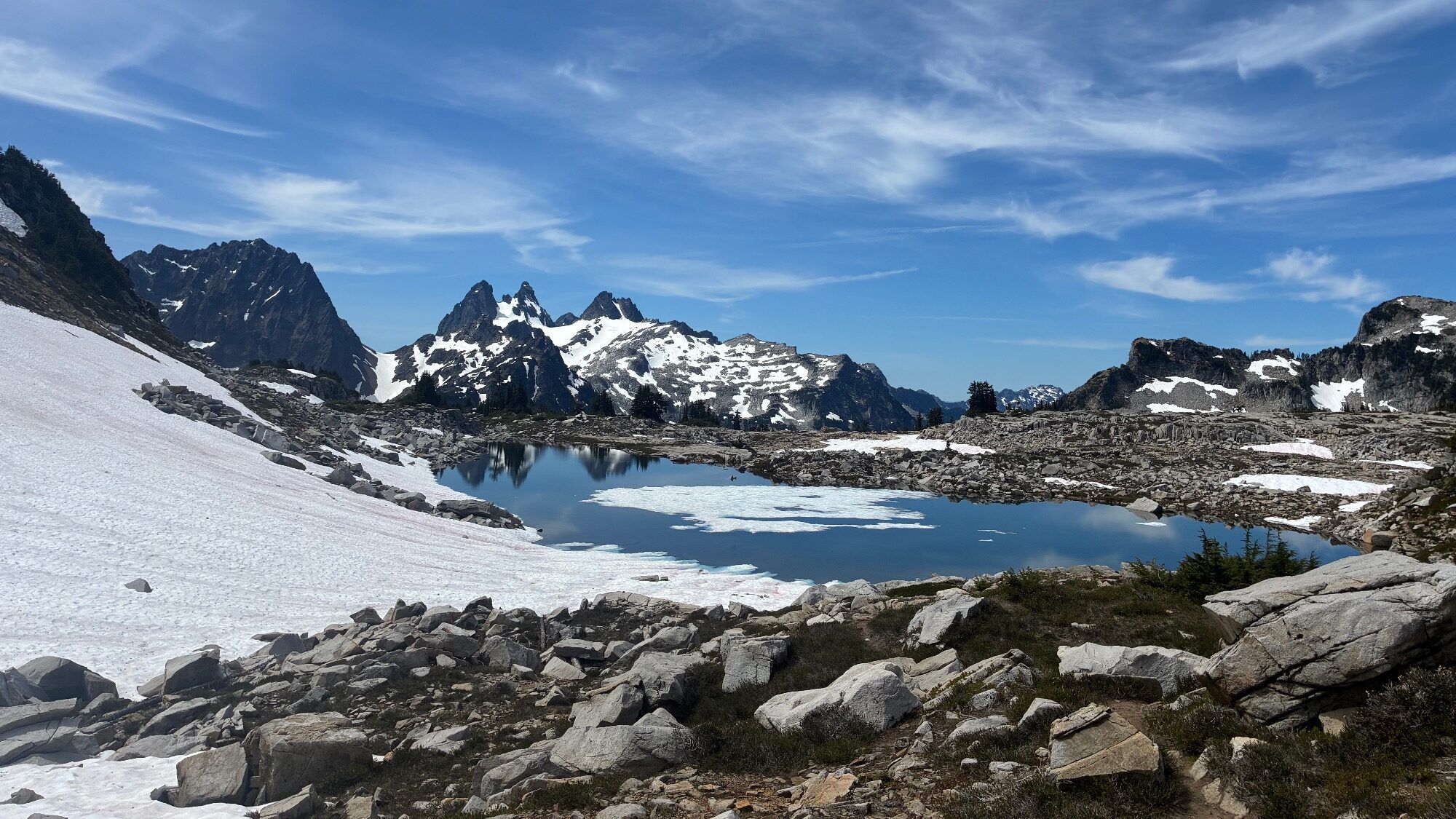 Rocky mountains with snow patches surround a clear alpine lake under a blue sky with wispy clouds.