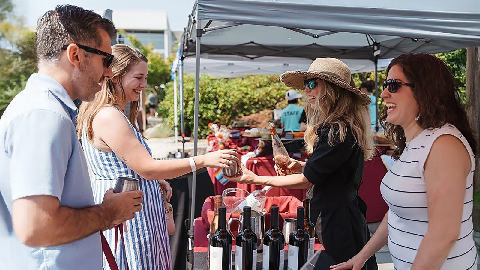 People at the South Lake District Block Party stand by a table with bottles of wine, smiling and interacting under a canopy on a sunny day—a perfect addition to your list of what to do in Seattle this week.