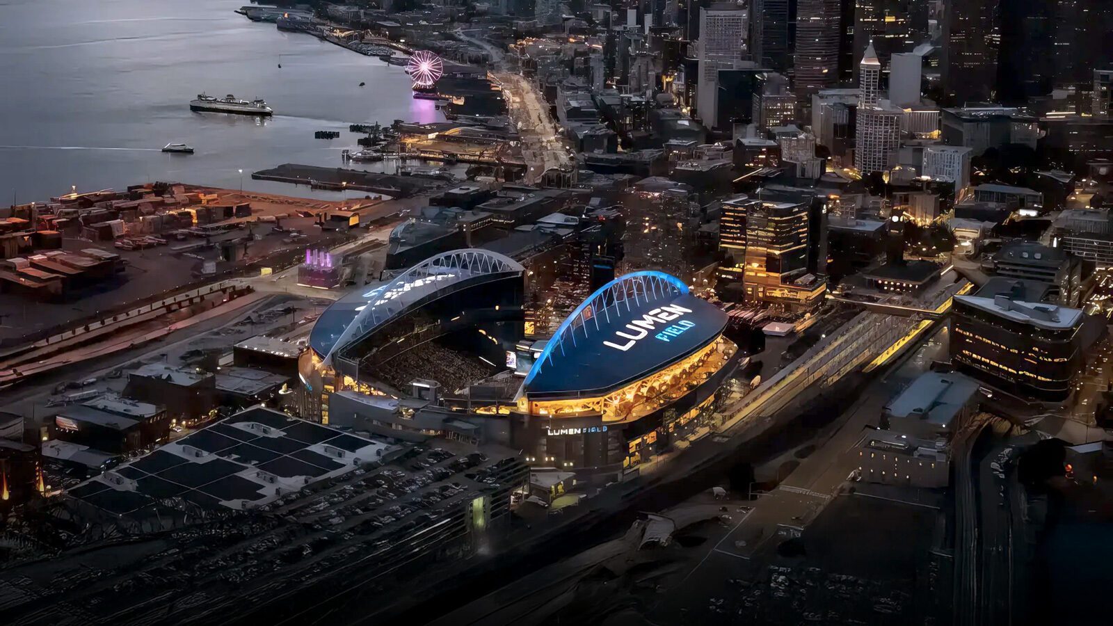 Aerial view of Lumen Field stadium illuminated at night in downtown Seattle, with adjacent waterfront, city buildings, and nearby streets visible.