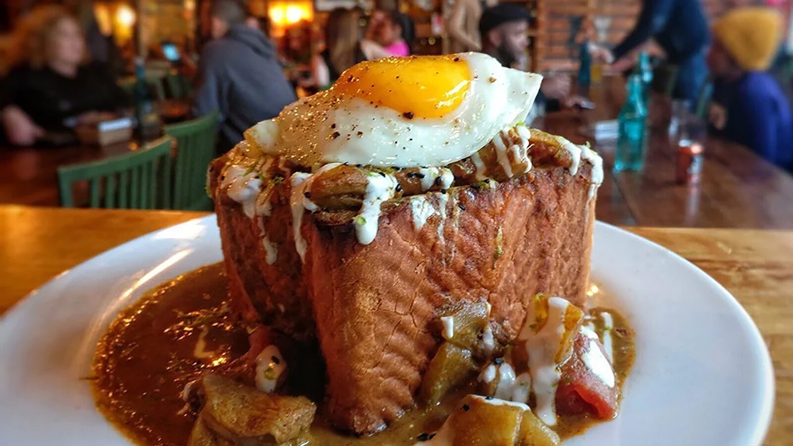 A thick slice of bread topped with fried potatoes, sausage gravy, and a sunny-side-up egg—this perfect September food is served on a white plate in a busy restaurant.